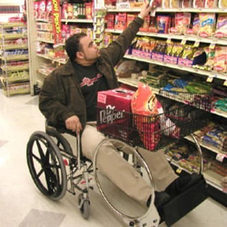 Man in Wheelchair Shopping Cart reaches for a product on a grocery store shelf.