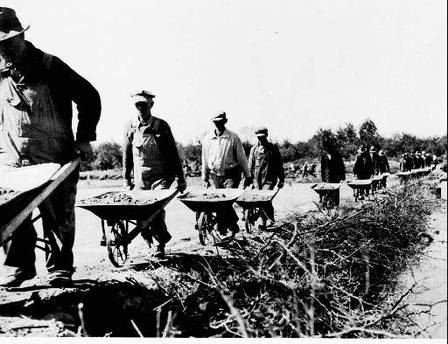 Men with wheelbarrows carry dirt in a line too far for the eye to see, along a river